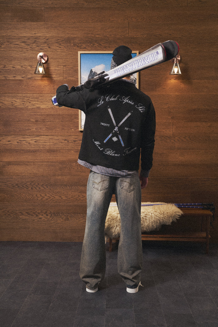 Person wearing a black long sleeved t-shirt with a logo, holding a skateboard, in a room with wooden walls and a bench.