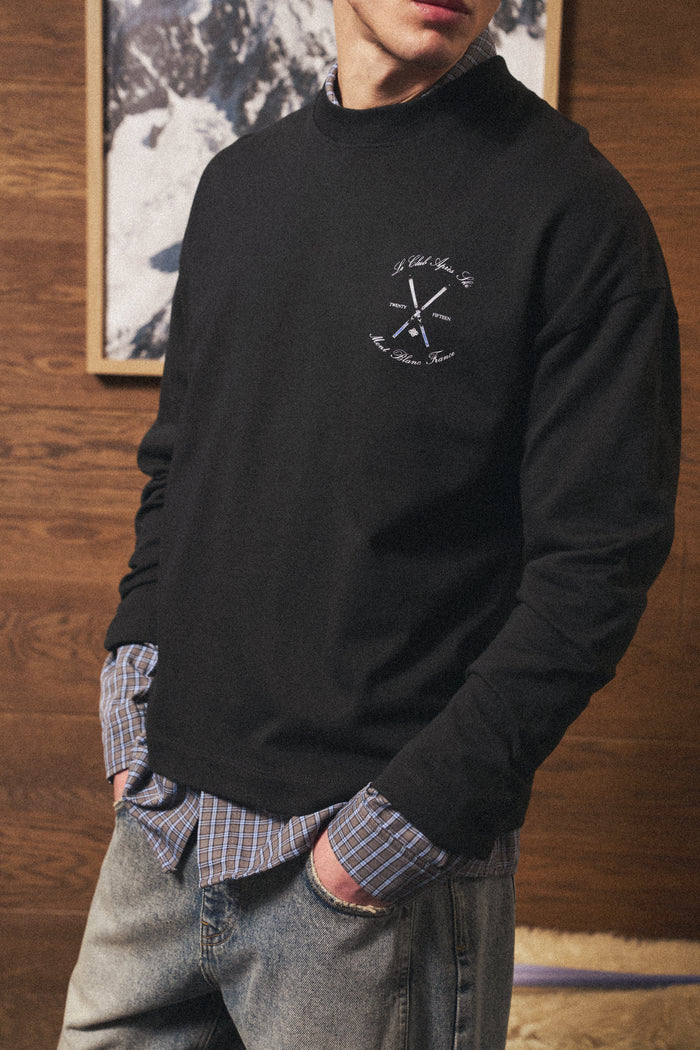 Man wearing a black long sleeved t-shirt with a logo, standing against a wooden wall.