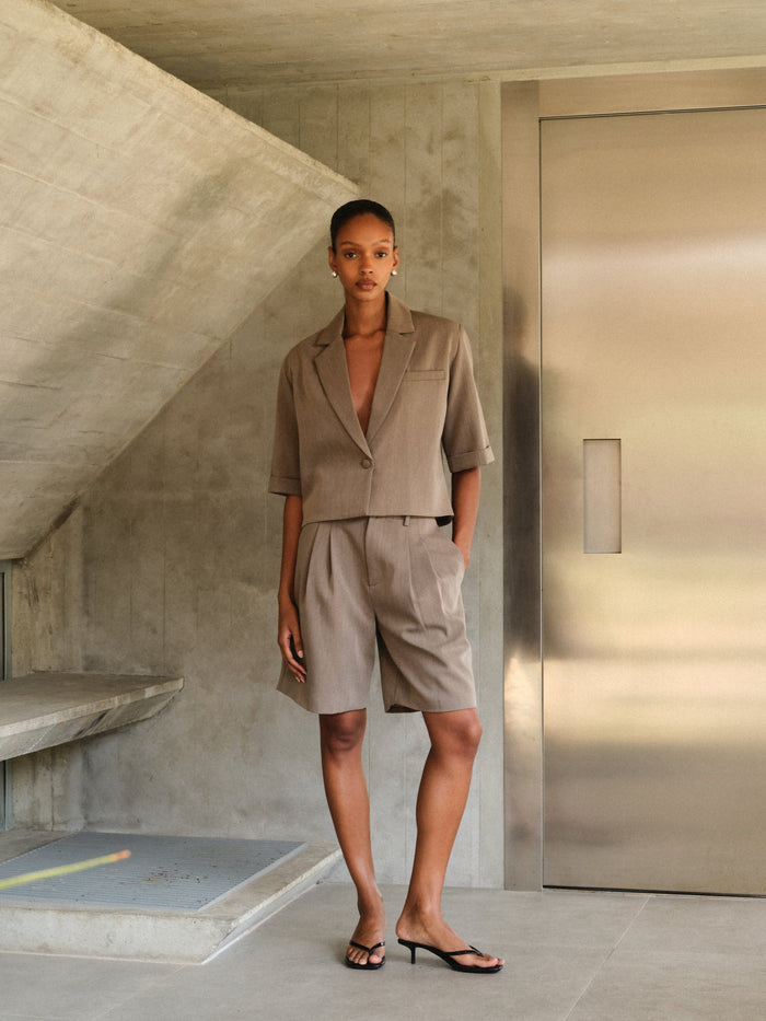 Woman in a beige tailored suit standing in a modern indoor setting with concrete walls and a metallic door.